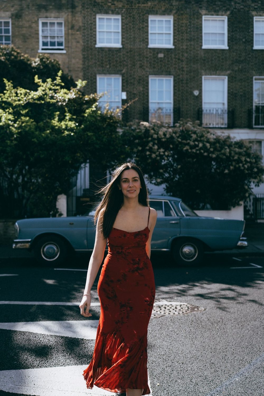Woman in a long red dress walking down a street of brick townhouses, a vintage car behind her, on a personal brand shoot