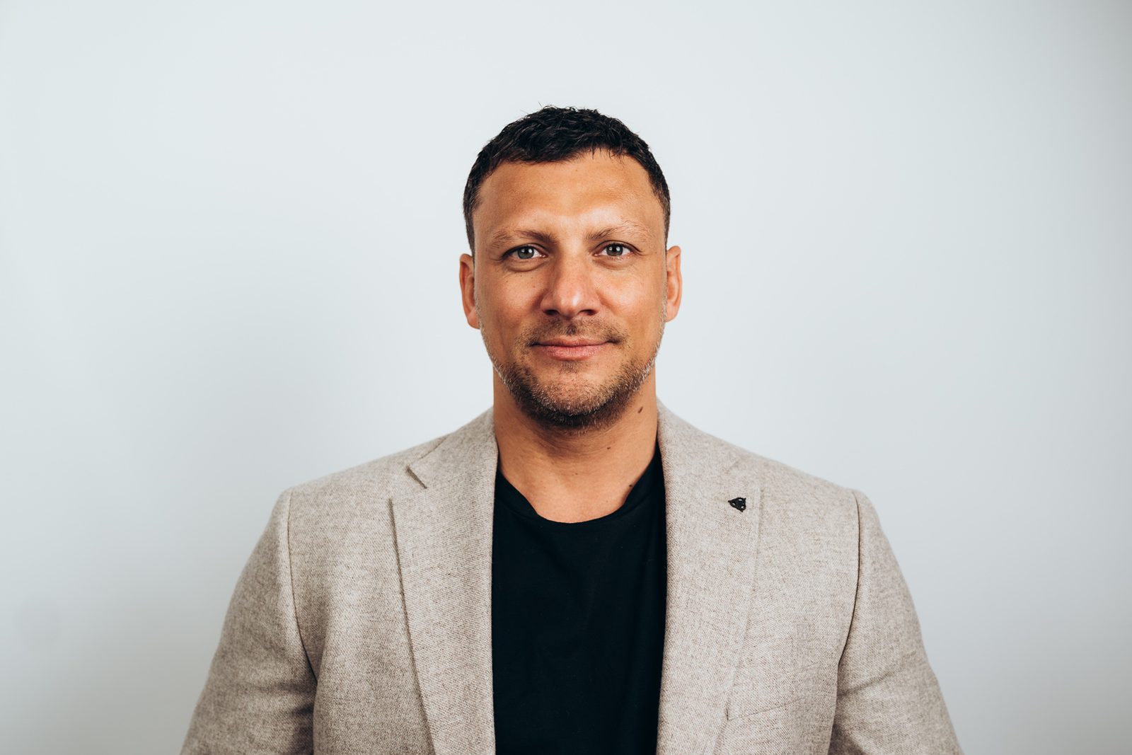 Headshot of a man in a beige blazer and black t-shirt against a plain pale backdrop